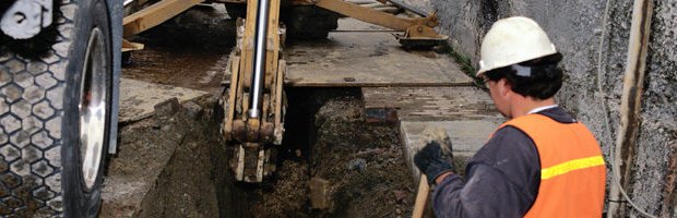 A worker guiding an excavator bucket into a utility trench