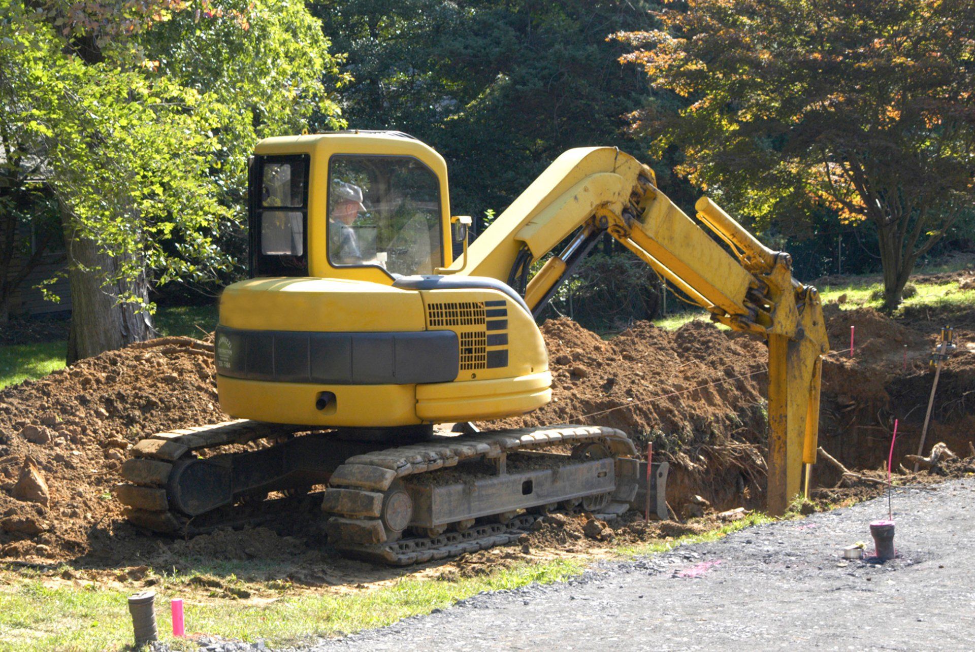 A compact yellow excavator digging a trench beside a gravel drive, with marking stakes in the ground