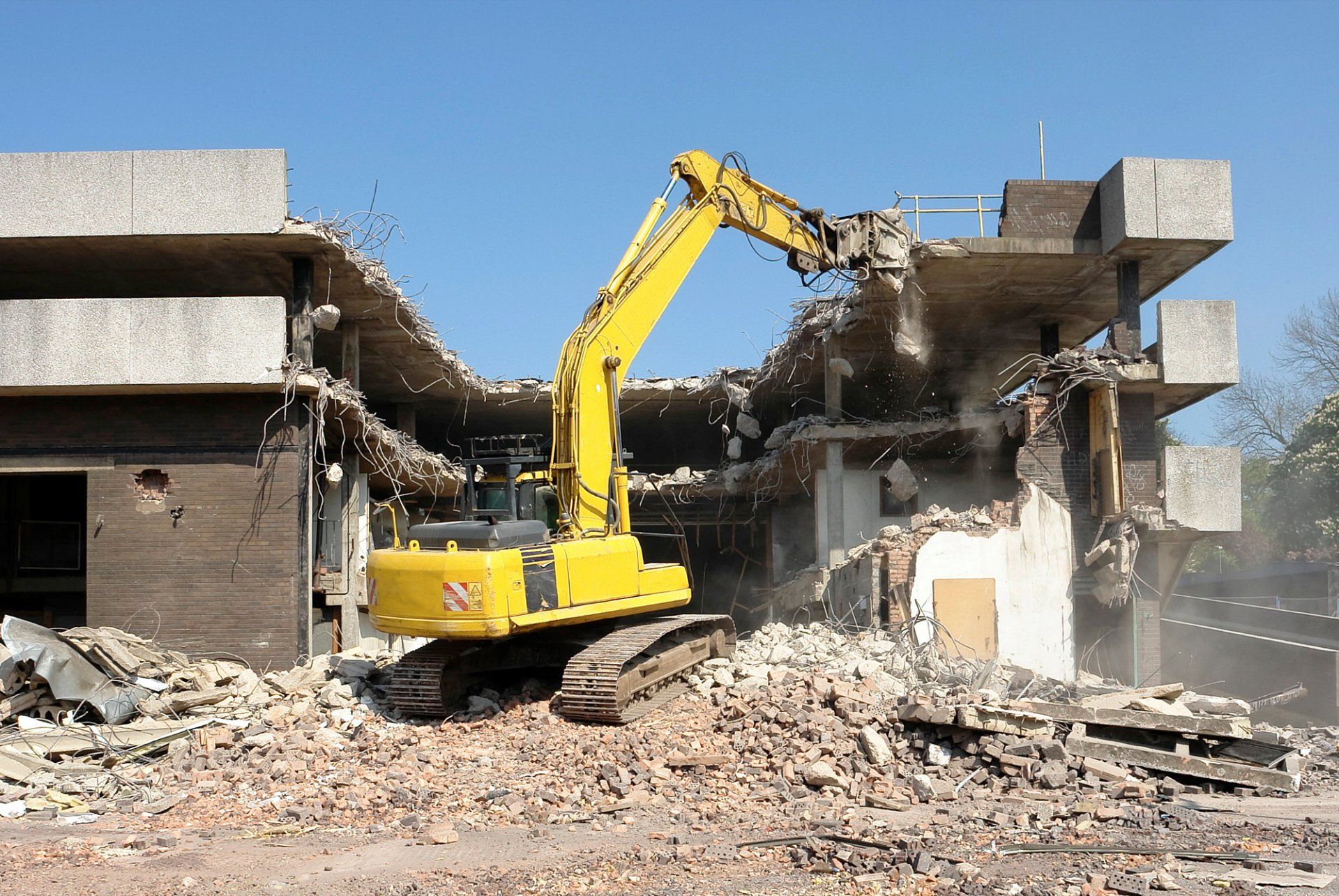 A yellow excavator tearing down a two-story concrete building, rubble piled in front